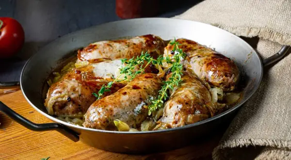 Pieczona w piekarniku biała kiełbasa surowa z cebulą i tymiankiem, podana w metalowej patelni.