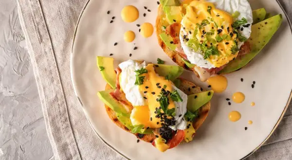 Två läckra eggs benedict på rostat bröd med avokado, bacon och hollandaisesås. Garnerade med persilja och svarta sesamfrön.