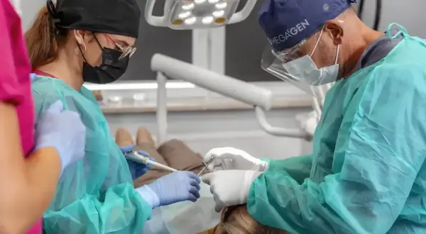 Dentysta i asystentka w maseczkach i fartuchach wykonują zabieg. Widać narzędzia i lampę zabiegową.