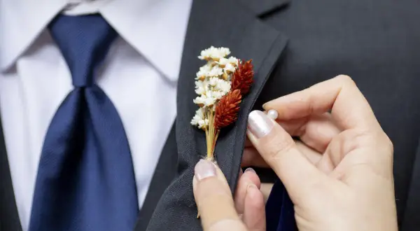 Dłoń przypina do marynarki mały bukiecik z suszonych kwiatów. To właśnie butonierka co to, ozdoba na specjalne okazje.