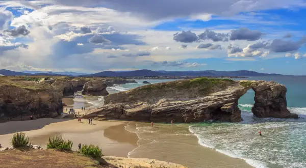 Espectacular arco natural en la playa de Esteiro, Ribadeo. Olas rompen en la arena dorada bajo un cielo nublado.