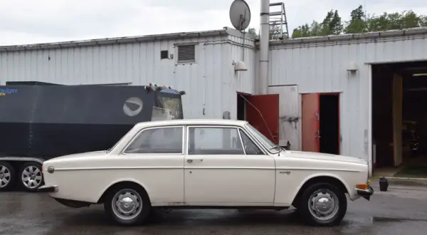 En vit Volvo 142 från 1973 står parkerad framför en industribyggnad.
