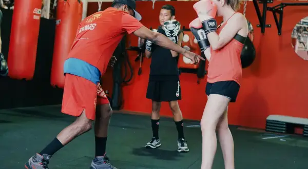 A boxing coach instructs a young woman on a proper duck punch. Another young person watches.