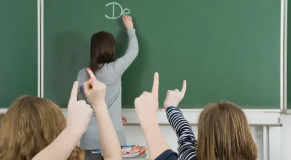 Lehrerin schreibt "De" an Tafel, Schüler heben Hände. Einblick in die bildungspolitik deutschland.