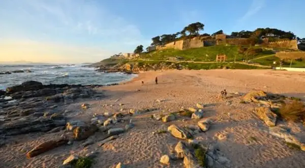 Atardecer en la playa concheira de Baiona. Olas rompen en la costa rocosa, mientras una fortaleza histórica corona la colina verde.