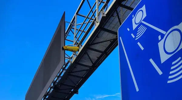 Żółta kamera na konstrukcji nad drogą, obok niebieskiego znaku z symbolami aparatów. To zapowiedź nowych odcinkowych pomiarów prędkości.