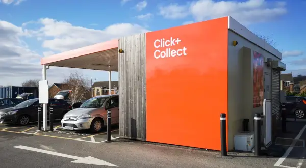 Zona de **click and collect** con coches aparcados bajo un toldo. Edificio naranja brillante con el texto "Click+ Collect".