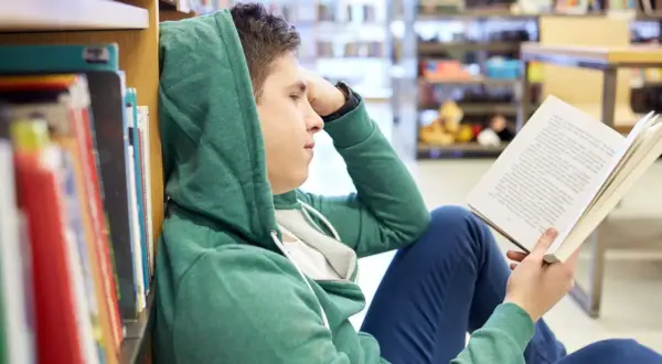 Chłopak w kapturze czyta książkę, szukając streszczenia makbeta. Siedzi na podłodze w bibliotece, opierając się o regał.