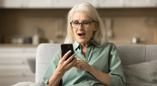 Starsza kobieta z zaskoczoną miną patrzy na telefon. Może właśnie dowiedziała się o przyznaniu zasiłku przedemerytalnego.