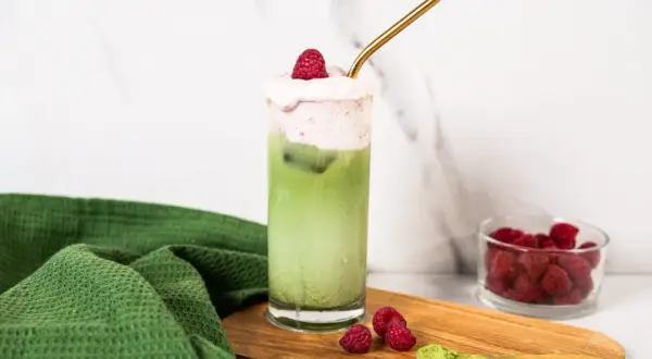 Orzeźwiające matcha z malinami, ozdobione pianką i świeżymi malinami, z dodatkiem proszku matcha na desce.
