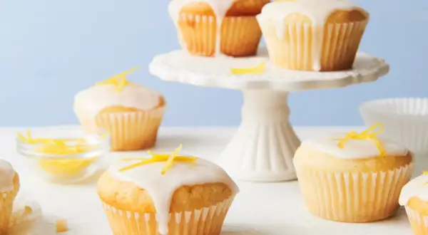Saftige vegane Zitronenmuffins mit Zuckerguss und Zitronenschale verziert, auf einem weißen Kuchenständer und Tisch.