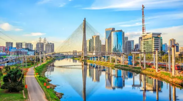 En bro med vajrar sträcker sig över en flod i São Paulo, med moderna skyskrapor som speglar sig i vattnet.