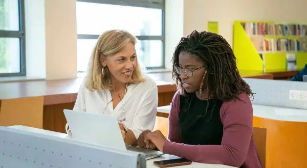 Dos mujeres en un aula, una rubia y otra afrodescendiente, revisan un portátil. La rubia sonríe, la otra parece preocupada, quizás porque su aptis caduca.