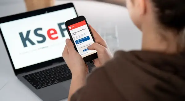 Osoba korzysta z telefonu, by dowiedzieć się, jak wystawić fakturę w KSeF. Na ekranie laptopa widnieje logo KSeF.