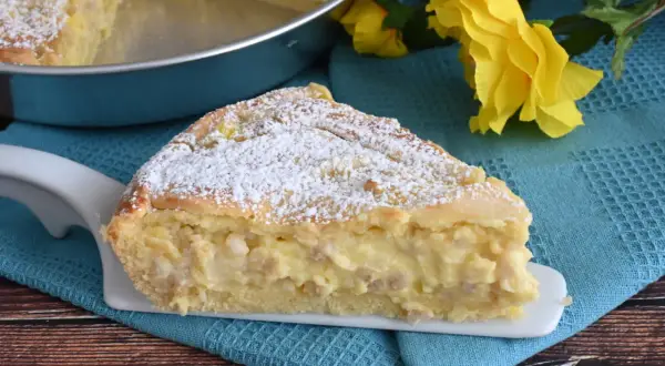 Una fetta di pastiera napoletana con crema pasticcera, cosparsa di zucchero a velo, su un piatto bianco, con fiori gialli sullo sfondo.