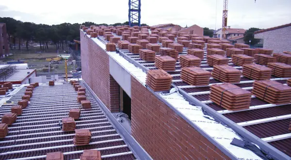 Pilotes de tejas rojas apiladas en la cubierta de una casa en construcción. Grúas en el fondo.
