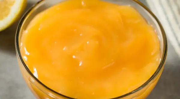 Gładki, żółty lemon curd w szklanej miseczce, obok połówka cytryny. Idealny do deserów.