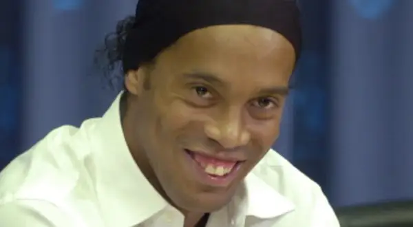 Uśmiechnięty Ronaldinho w białej koszuli i czarnej opasce na głowie, wspominający swoje najlepsze lata w różnych klubach.