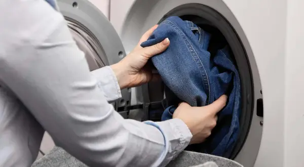 Hände legen Jeans in die Waschmaschine. Das passende Jeans waschen Programm wird ausgewählt.