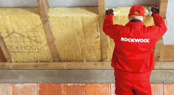 Pracownik w czerwonym stroju Rockwool montuje wełnę mineralną, wykonując ocieplenie murłaty od wewnątrz.
