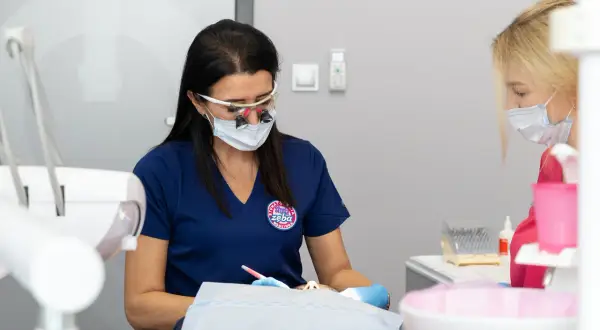 Dentystka w okularach powiększających i masce pracuje nad pacjentem, który odczuwa drętwienie po znieczuleniu zęba.