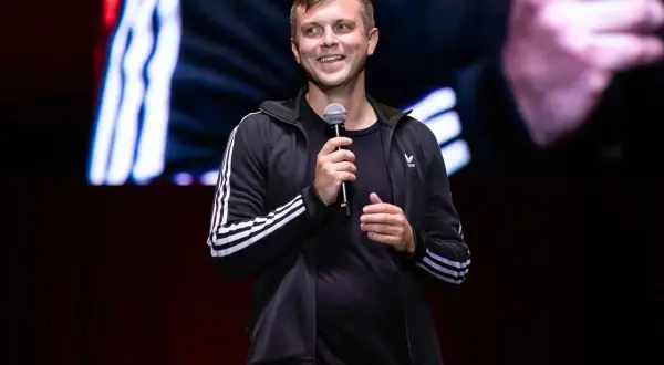 Uśmiechnięty Krajewski na scenie, trzyma mikrofon. Jego stand up rozgrzewa publiczność.