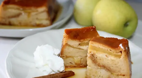 Delicioso pudin de manzana con caramelo, crema batida y canela. Dos porciones servidas en un plato blanco, con dos manzanas verdes al fondo.