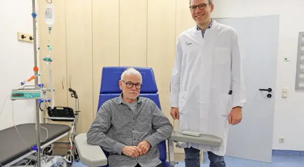 Ein Arzt steht neben einem Patienten, der für eine Alzheimer Behandlung in einem Behandlungsstuhl sitzt. Infusionsständer und medizinische Geräte sind im Hintergrund sichtbar.