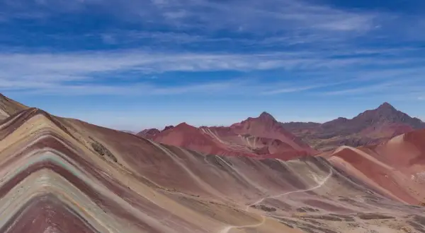 La montagne Vinicunca, aux couleurs vives, s'étend sous un ciel bleu parsemé de nuages. Un sentier serpente à travers ses flancs striés.