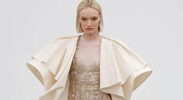 Modelka w kremowym, falbaniastym płaszczu i zdobionej sukni prezentuje dzieło haute couture.