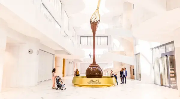 Imponerande chokladfontän i Lindt Home of Chocolate-museet. Familjer beundrar den gigantiska skulpturen i den ljusa, moderna byggnaden.