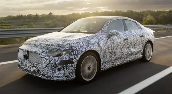 Zamaskowany prototypowy Mercedes CLA 2025 na drodze, z widokiem na zachodzące słońce i drzewa w tle.