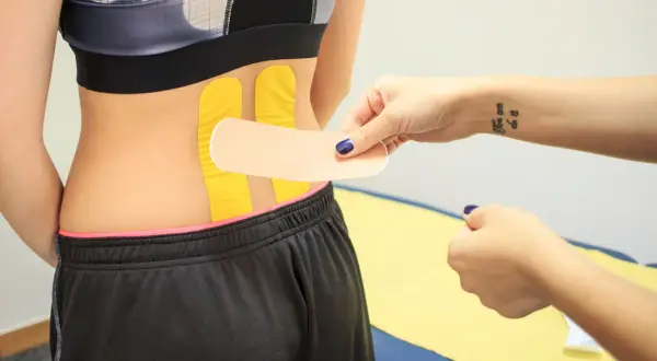 Naklejanie tejpy na plecy w celu wsparcia mięśni. Dłoń z tatuażem "You can do it" przygotowuje plaster.
