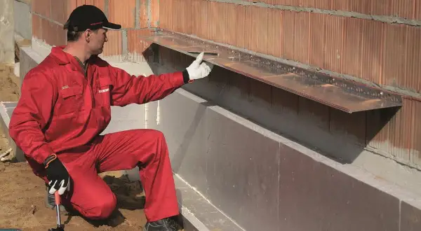 Pracownik w czerwonym kombinezonie montuje okapnik na ścianie zewnętrznej, przygotowując ją do montażu wełny mineralnej.