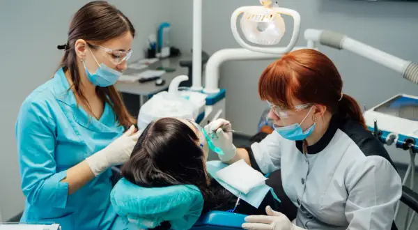 Dwie kobiety w maseczkach i okularach ochronnych pracują przy pacjencie w gabinecie stomatologicznym. Jedna z nich, asystentka stomatologiczna, podaje narzędzia.