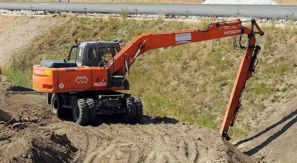 Pomarańczowa koparka Hitachi Zaxis 160W pracuje na budowie. Operator posiada odpowiednie uprawnienia na koparki, by sprawnie wykonywać prace ziemne.