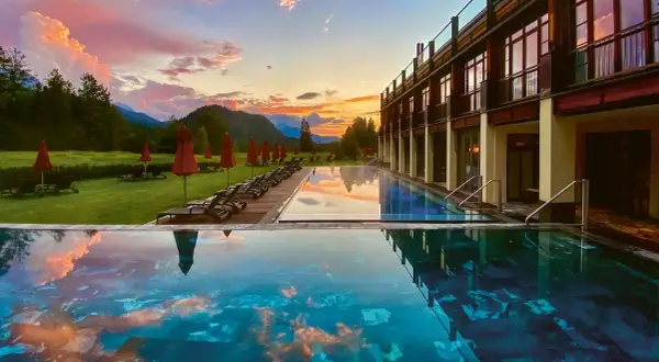 Abendrot über den Alpen spiegelt sich im Infinity-Pool eines der besonderen Hotels Bayern. Liegestühle und rote Sonnenschirme säumen den Rasen.