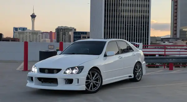 Biała Toyota Altezza z tuningowym body kitem i spojlerem na dachu parkingu, w tle wieżowce i wieża widokowa.