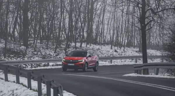 Czerwony SUV jedzie po mokrej drodze, otoczonej ośnieżonym lasem. Kierowca uważa, bo to może być gololedz.