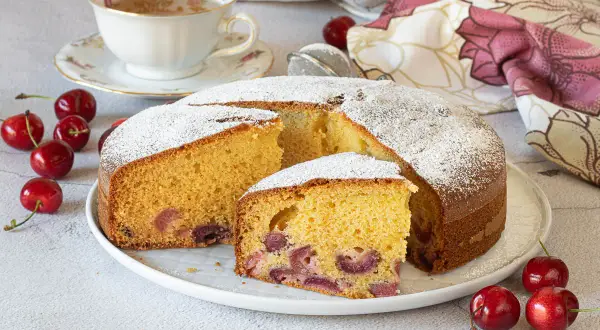 Una fetta di torta alle ciliegie, soffice e profumata, cosparsa di zucchero a velo. Ciliegie fresche e una tazza di tè completano la scena.