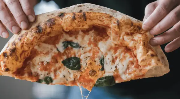 Hände halten ein Stück Pizza mit luftigem Rand, geschmolzenem Käse und Basilikum.