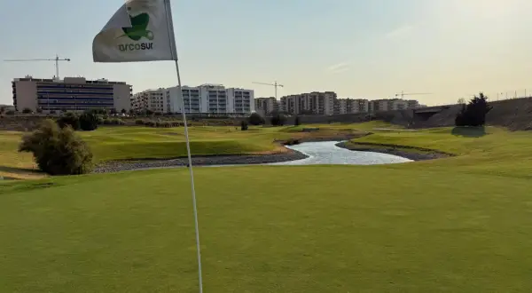Bandera de Arcosur en un campo de golf, con edificios residenciales y grúas de construcción al fondo.