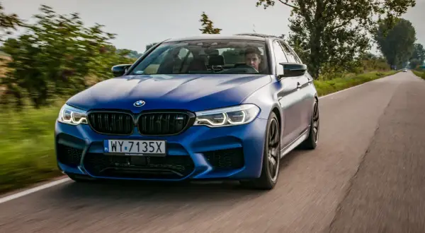 Niebieskie BMW M5 F90 pędzi po wiejskiej drodze, kierowca skupiony na trasie.
