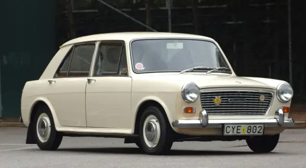 En krämvit Austin 1100 står parkerad på asfalt. Bilen har en klassisk design med kromade detaljer och en tydlig grill.