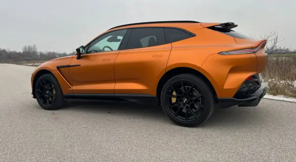 Pomarańczowy Aston Martin DBX z czarnymi felgami i przyciemnianymi szybami na tle pochmurnego nieba.