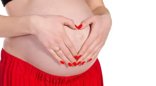 Mani con unghie rosse formano un cuore sul pancione. Si possono fare le unghie in gravidanza, con cura e attenzione.