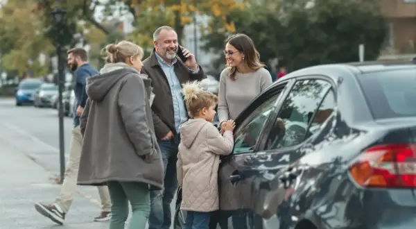 Familie am Auto, Vater telefoniert, vielleicht wegen eines Problems, das die Unfallkasse des Bundes betrifft.