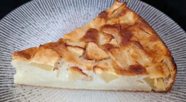 Tranche de clafoutis aux pommes dorée et moelleuse, garnie de morceaux de fruits fondants, sur une assiette texturée.