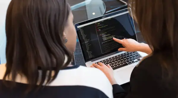 Dwie kobiety analizują kod na laptopie. Jedna wskazuje palcem na ekran, druga pisze. Tak wygląda praca programisty.