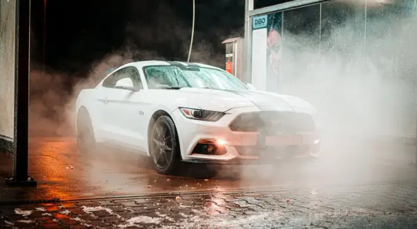 Biały Mustang w obłokach pary podczas mycia aut. Woda spływa po karoserii, tworząc efektowną mgiełkę.